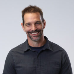 Justin - An owner at Community Real Estate. A smiling white man, with brown hair and salt and and pepper beard, wearing a dark blue tshirt, underneath a black button up shirt.